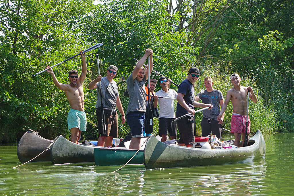 Eine Gruppe junger Menschen steht jubelnd mit erhobenen Paddeln in mehreren Kanus auf einem Fluss
