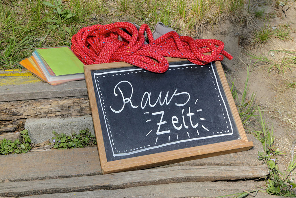 Eine Schiefertafel mit der Aufschrift „RausZeit“ liegt mit farbigen Karten und einem roten Seil auf Holzbohlen im Freien